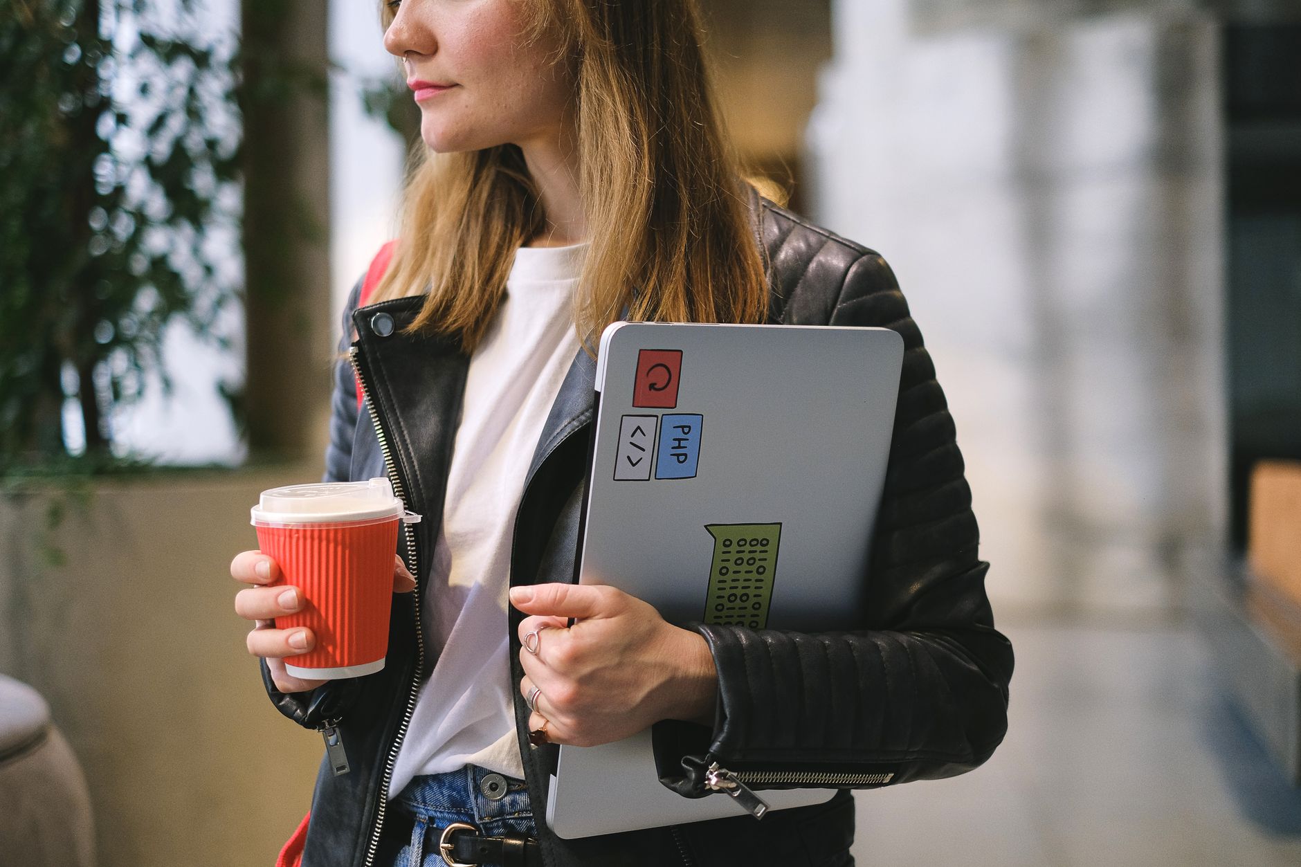 Jeune femme en veste de cuir noire tenant un ordinateur portable et une tasse de café à emporter à l'intérieur.