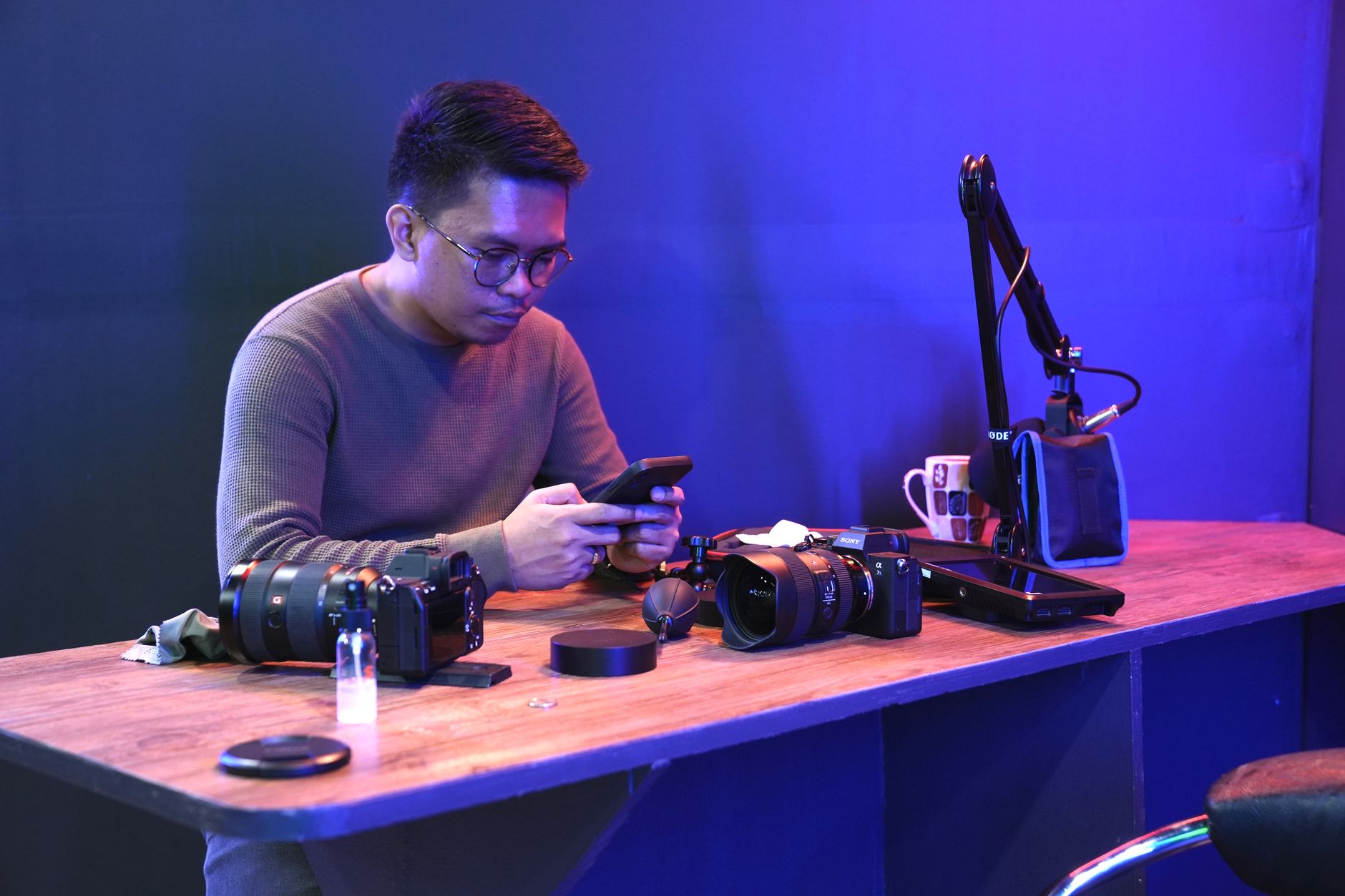 Homme assis à un bureau avec des caméras et un microphone, utilisant un smartphone.