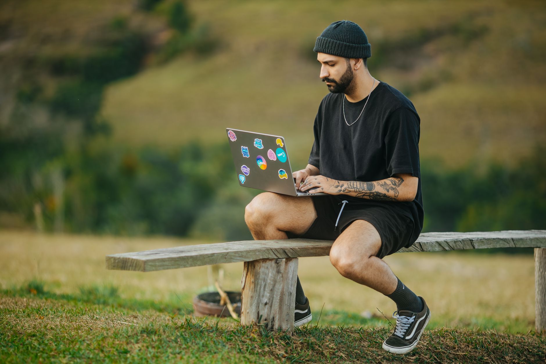 Homme décontracté avec barbe et ordinateur portable assis à l'extérieur, concentré sur le travail dans un cadre rural paisible.