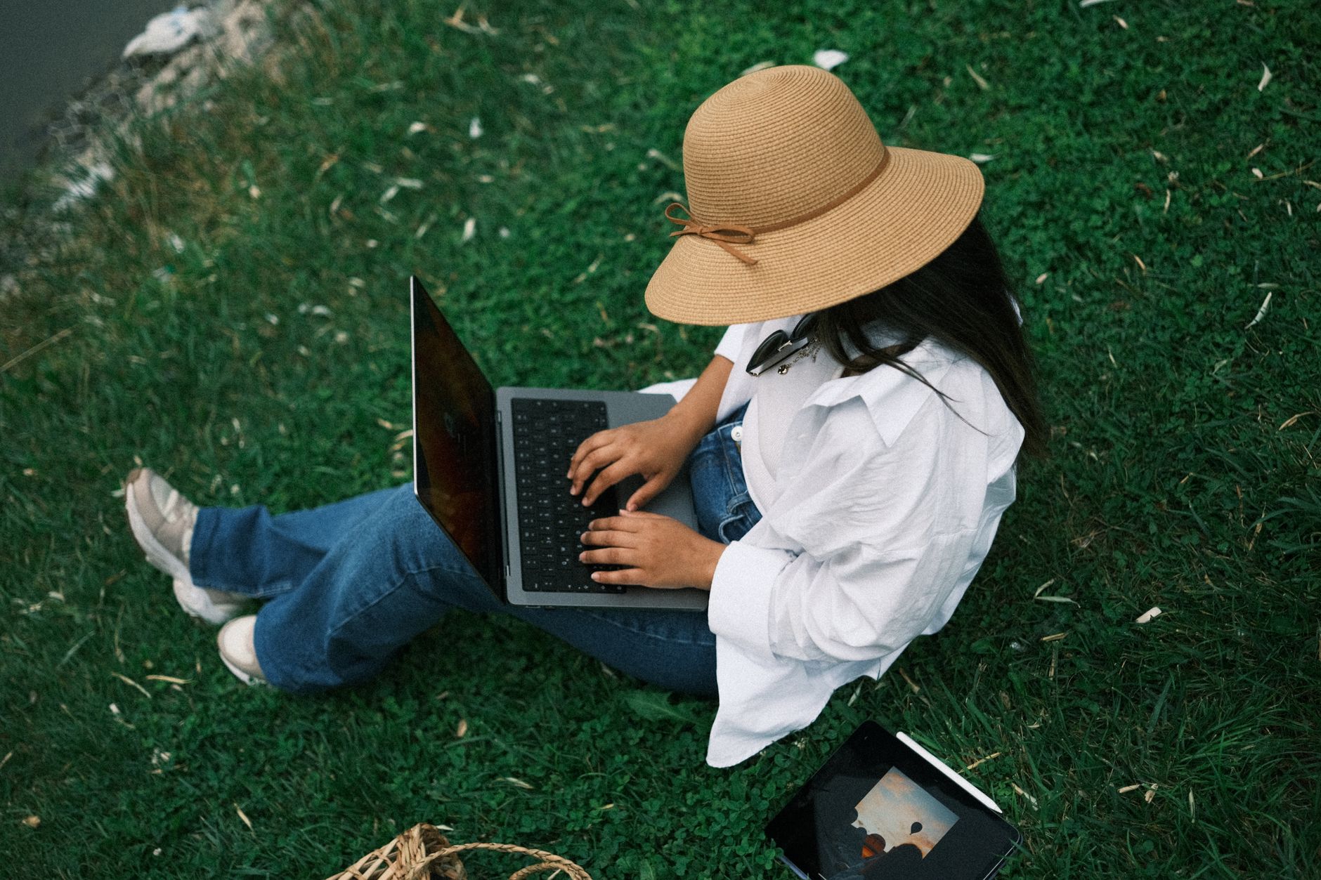 Femme assise dans un parc avec un ordinateur portable, portant un chapeau et travaillant à distance en plein air.