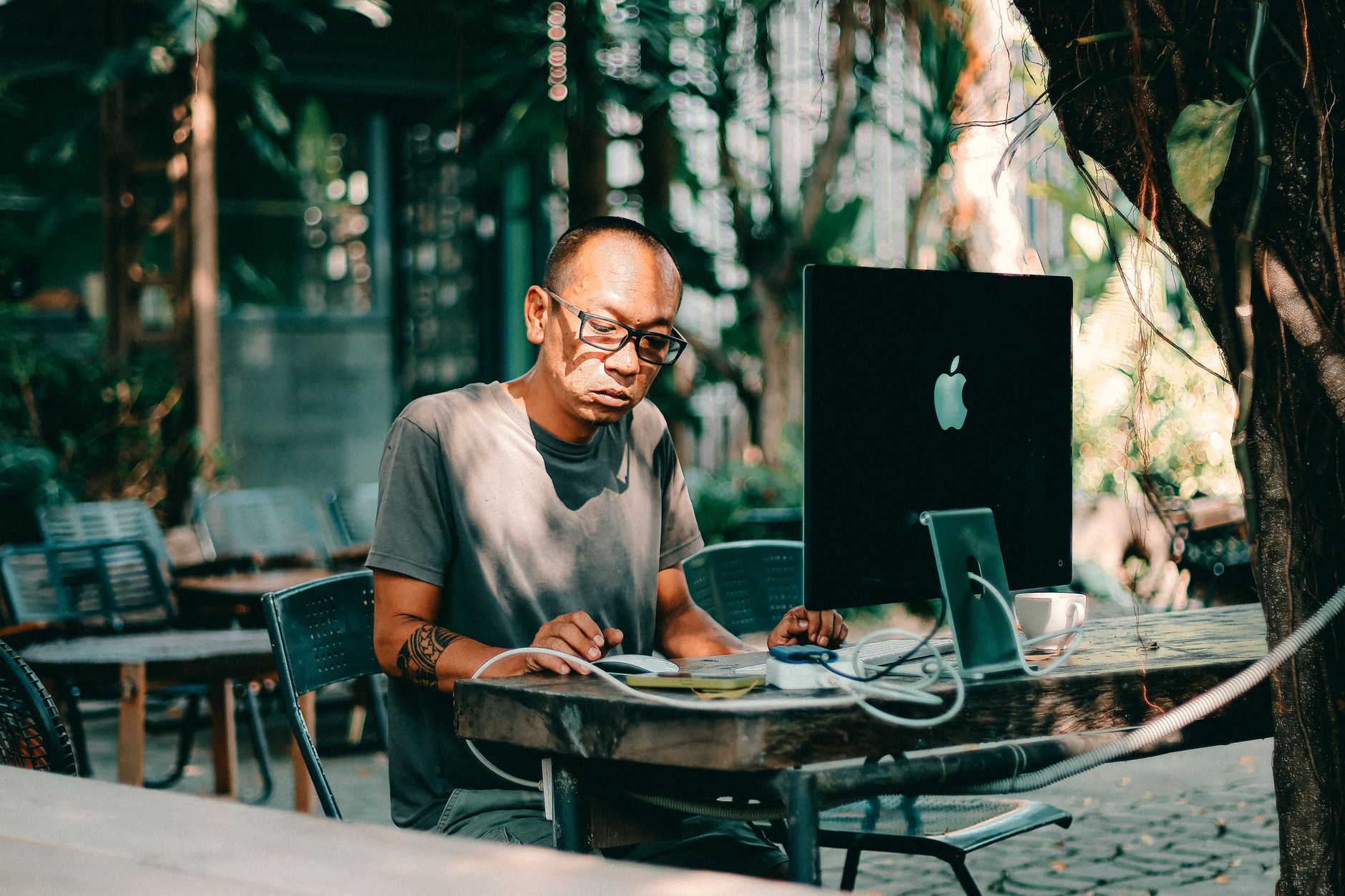 Homme adulte avec des lunettes travaillant sur un ordinateur à l'extérieur dans un café ensoleillé.