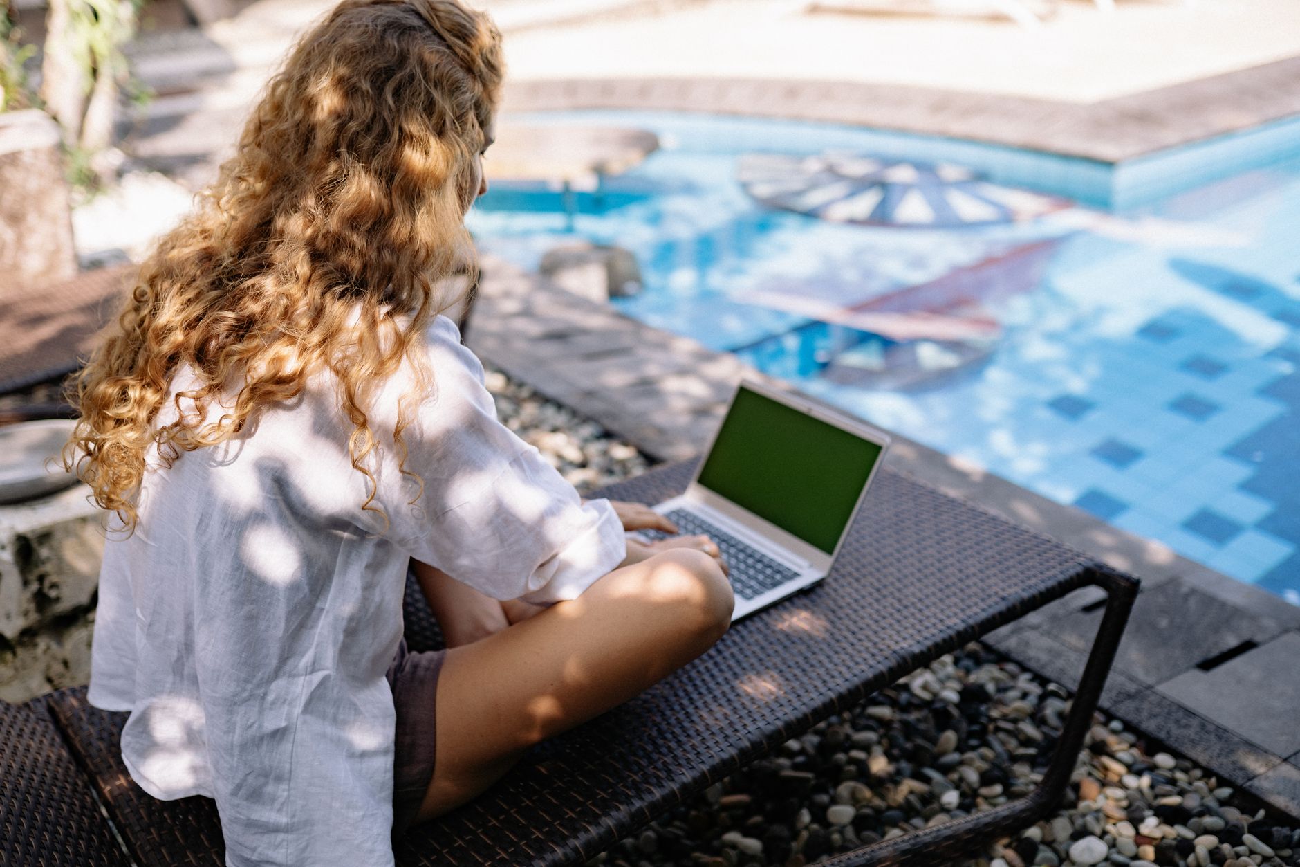 Femme aux cheveux bouclés travaillant sur un ordinateur portable à l'extérieur près d'une piscine pendant l'été, incarnant un style de vie de nomade numérique.