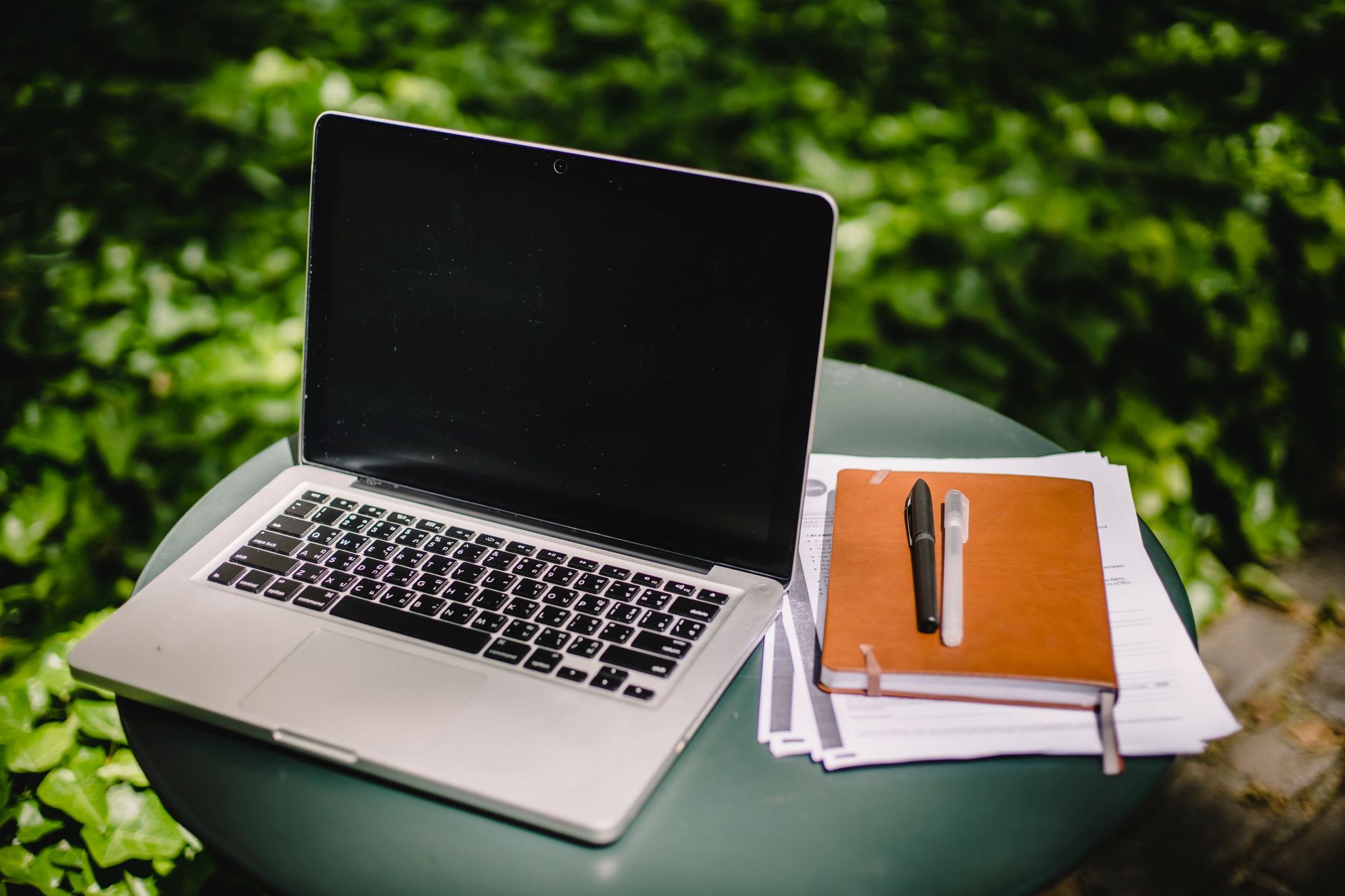Ordinateur portable moderne avec écran vide posé près d'un cahier marron et des papiers sur une table ronde contre un jardin vert flou